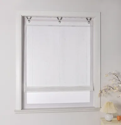 A white roller blind with a subtle pattern is installed in a window frame. A small lamp and some decorative branches are visible on the right side.