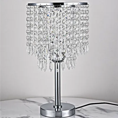 A crystal table lamp with a chrome base and stem is shown against a light gray background, resting on a marble-like surface.