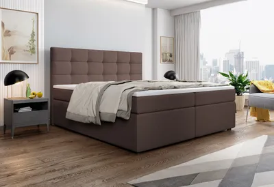 A bedroom scene with a large brown boxspring bed as the focal point, set against a backdrop of a window with a city view and a nightstand with a lamp.