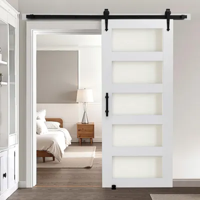 A white sliding barn door with frosted glass panels is open, revealing a bedroom with a bed, nightstand, and lamp.