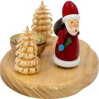 Ein hölzerner Kerzenhalter zeigt einen kleinen Weihnachtsmann in rotem Gewand mit einem grünen Zweig in der Hand neben zwei kunstvoll geschnitzten Bäumen auf einer runden Holzplatte