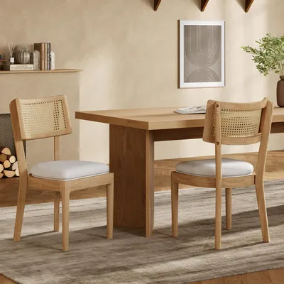 Two dining chairs with rattan backs and light-colored cushions are placed around a wooden dining table in a room with a fireplace and a framed picture on the wall.