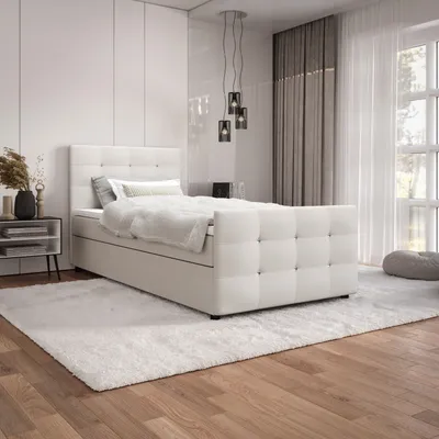 A bedroom scene with a white boxspring bed as the main focus, set against a white wall and wooden floor.