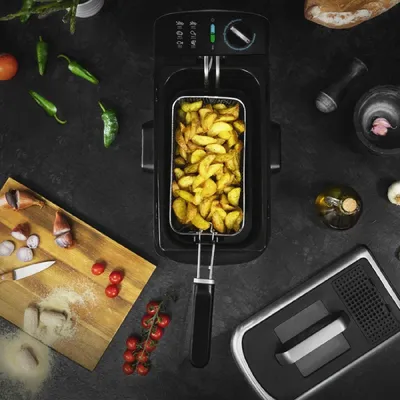 A deep fryer filled with french fries, surrounded by ingredients like tomatoes, peppers, and garlic, on a dark surface.