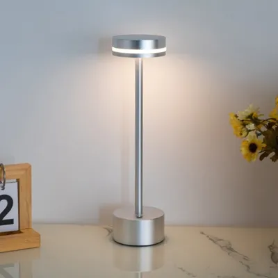 A modern LED table lamp with a silver finish is displayed on a reflective surface, with a blurred background and a small wooden calendar on the left.
