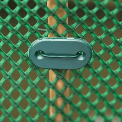A close-up of a green plastic clip attached to a green mesh fence with a wooden post behind it.
