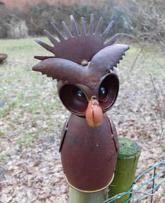 A rusty metal owl garden decoration is mounted on a wooden post in an outdoor setting with dry leaves on the ground.
