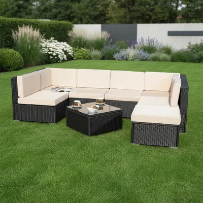 A black polyrattan lounge set with beige cushions is arranged on a green lawn in a garden setting, with a wall and plants in the background.