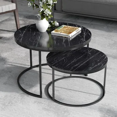 A set of two round nesting coffee tables with a black marble-like top and black metal legs, placed on a light gray floor. A white vase with white flowers and a book are on the larger table.