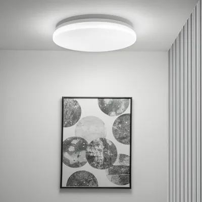 A modern white round LED ceiling light is mounted on a white ceiling above a framed abstract artwork with circular patterns.