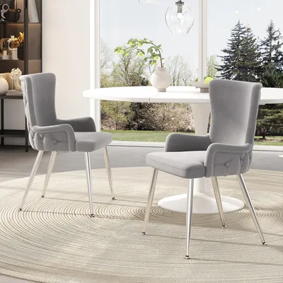 Two gray velvet dining chairs with silver metal legs are placed around a white round dining table in a well-lit room with a large window overlooking a garden.
