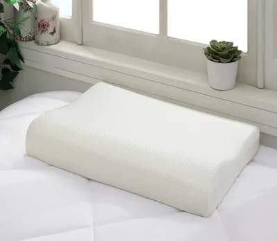 A white orthopedic pillow is placed on a white quilted surface, with a window and decorative items in the background.