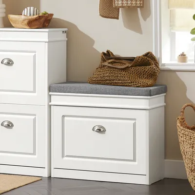 A white shoe bench with a gray cushion and a woven bag on top, placed next to another white storage unit.