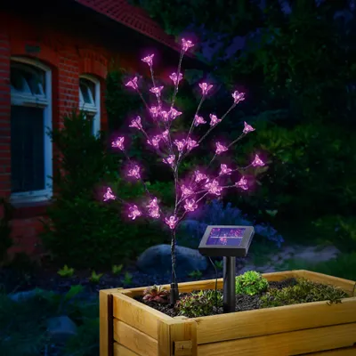 A pink LED blossom tree with a solar panel is planted in a wooden planter box next to a brick house at night.