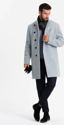 A man wearing a gray coat, blue sweater, and black pants stands against a white background.