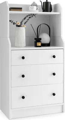 A white dresser with three drawers and a shelf on top, decorated with a vase of flowers and a small lamp.