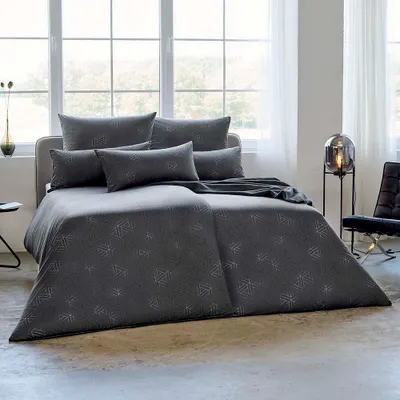 A neatly made bed with gray bedding and several pillows in a well-lit room with large windows.
