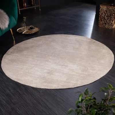 A round beige rug is placed on a dark wooden floor in a living room setting. A green sofa and a gold side table are visible in the background.