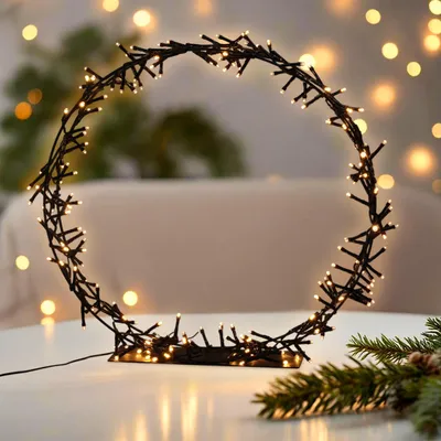A circular LED light decoration with a stand, placed on a table with a blurred background of lights and a hint of a Christmas tree branch.