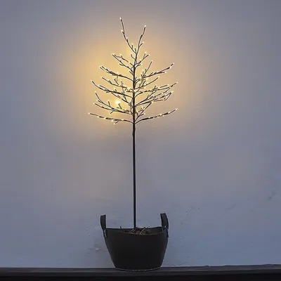 A decorative LED tree with warm white lights in a black pot, set against a light gray wall.