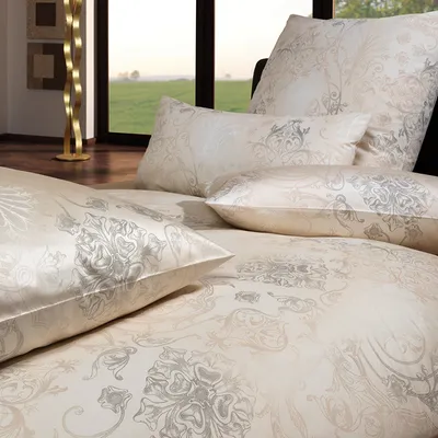 A close-up view of a bed with patterned bedding and pillows, with a window and a lamp in the background.