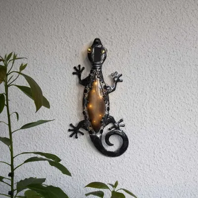 A decorative solar-powered gecko light is mounted on a textured wall, with a plant in the foreground.