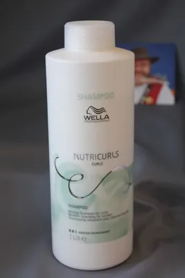 A white bottle of Wella Professionals Nutricurls shampoo is displayed on a gray fabric background. A small, out-of-focus image of a person in traditional attire is visible in the background.
