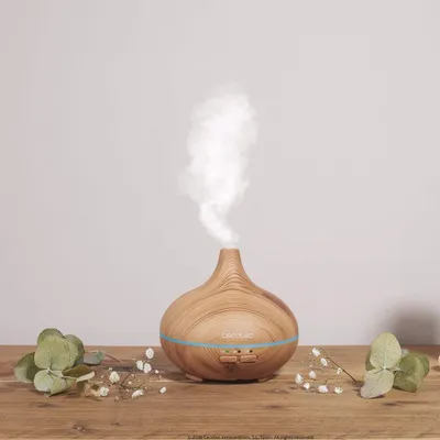 A wooden essential oil diffuser emitting mist, placed on a wooden surface with greenery and small white flowers around it.