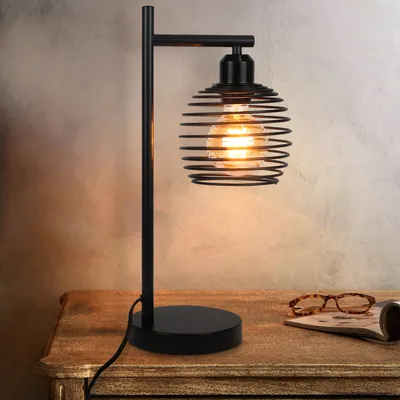 A black metal table lamp with a spiral cage shade sits on a wooden table.