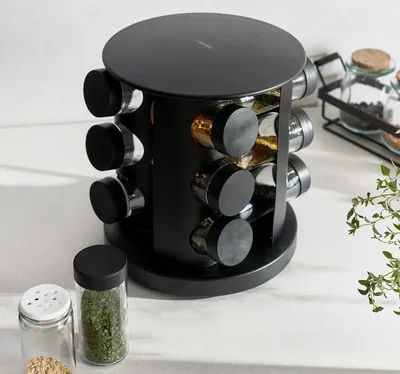 A black rotating spice rack with 12 glass jars filled with spices, placed on a white countertop.