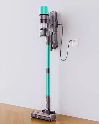 A teal and gray cordless vacuum cleaner is mounted on a charging station attached to a white wall. The vacuum is standing upright, with its head resting on a wooden floor.