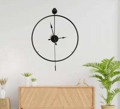 A modern black metal wall clock with a minimalist design hangs on a white wall above a wooden cabinet.