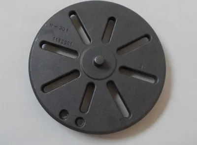 A round, dark gray metal grate with slots is shown on a light gray surface.