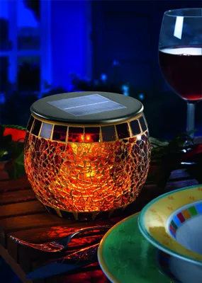 A solar-powered mosaic LED table lamp is displayed on a wooden table, set for a meal with a glass of red wine and plates.