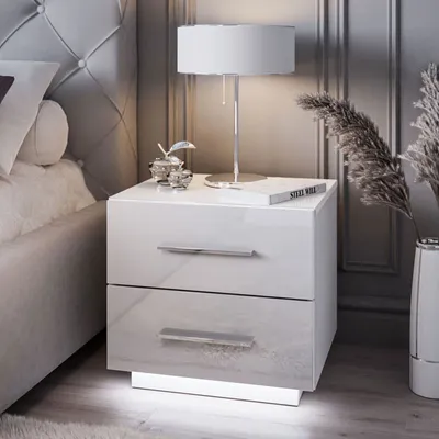 A modern white nightstand with two drawers is placed next to a bed, illuminated by subtle LED lighting at its base. A table lamp sits on top, alongside decorative items and a book titled "STEEL WALL."