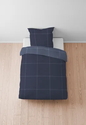 A neatly made bed with a blue plaid duvet cover and pillow on a wooden floor.