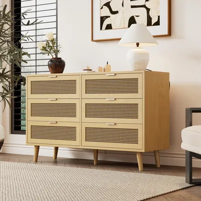 A wooden dresser with rattan woven drawer fronts sits in a well-lit room, next to a window and a piece of abstract art.