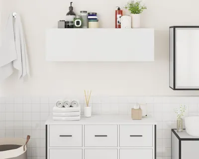 A clean, minimalist bathroom scene featuring a white wall-mounted cabinet above a white vanity with drawers. Several rolled towels and a reed diffuser are on the vanity. A towel hangs on the left.