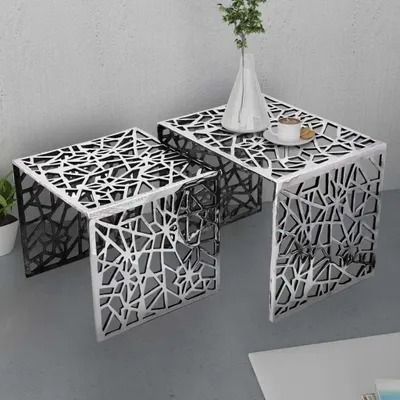Two nesting side tables with a geometric cut-out pattern are shown in a modern interior setting. One table has a cup of coffee and a cookie on it, and a vase with greenery is placed on the larger table.