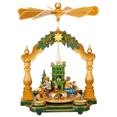 Das Bild zeigt eine weihnachtliche Pyramide mit einer Szene in Großmutters Stube wo Figuren um einen grünen Ofen versammelt sind und ein goldener Propeller obenauf thront während Tannenzweige und ein Stern die Szenerie schmücken