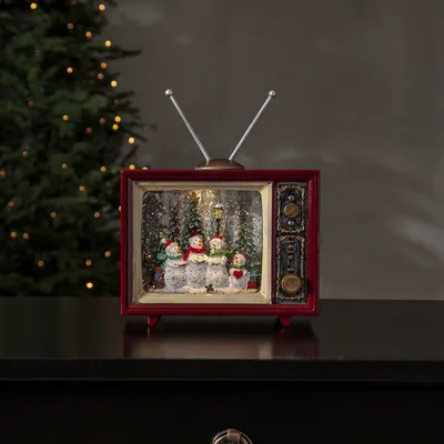 A decorative retro television with a Christmas scene inside, placed on a dark surface with a Christmas tree in the background.