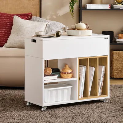 A white rolling storage cabinet with wooden dividers is placed on a brown carpet next to a sofa with pillows.