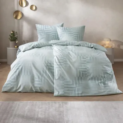 A neatly made bed with light blue bedding and pillows, set in a softly lit room with decorative wall art and a small Christmas tree.