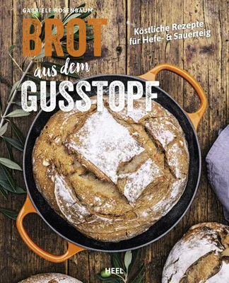 A rustic overhead shot of a loaf of bread in a cast iron pot, surrounded by olive branches and other loaves of bread on a wooden surface.