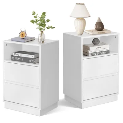 Two white bedside tables are shown side-by-side against a white background. The table on the left has a book, a clock, and a vase with greenery on top, and two drawers below. The table on the right has a lamp, a vase, and books on top, and two drawers belo