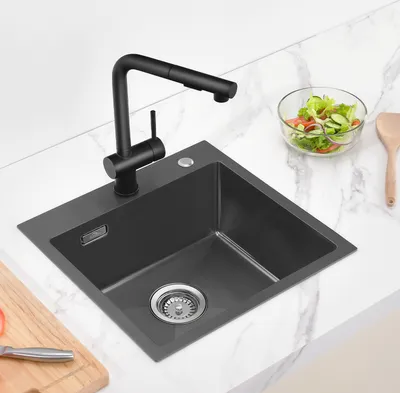 A dark gray kitchen sink with a black faucet is shown on a white marble countertop. A bowl of salad and a wooden spoon are visible to the right, and a cutting board with a knife is to the left.