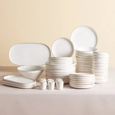 A collection of white porcelain dinnerware with gold trim, arranged on a light pink surface against a light beige background.