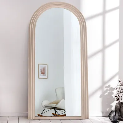 A full-length arched mirror with a light wood frame, leaning against a light-colored wall. A white rocking chair is reflected in the mirror.
