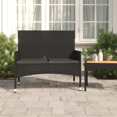 A black poly rattan garden bench with cushions is shown on a paved patio in front of a house with large windows.