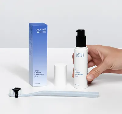 A hand holds a bottle of Alpine White Colour Corrector Serum next to its box and a toothbrush with black paste on it.